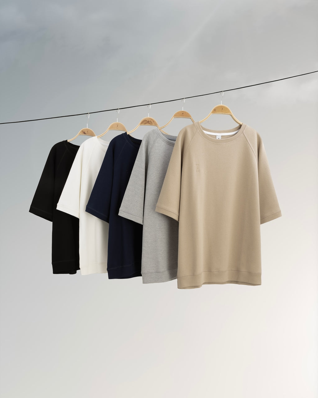 Five shirts in black, white, navy, gray, and beige hanging on a line against a light gray background.