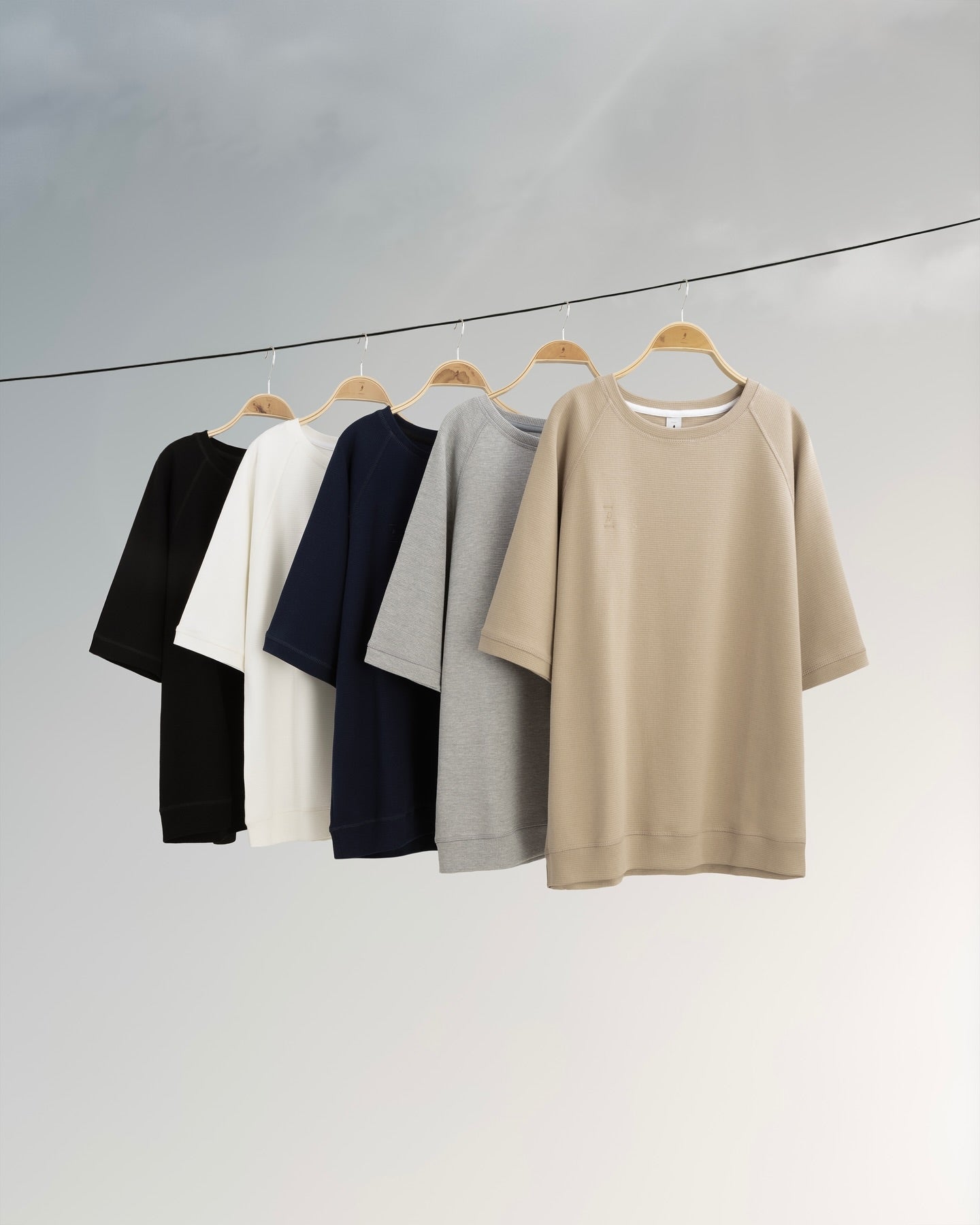 Five shirts in black, white, navy, gray, and beige hanging on a line against a light gray background.
